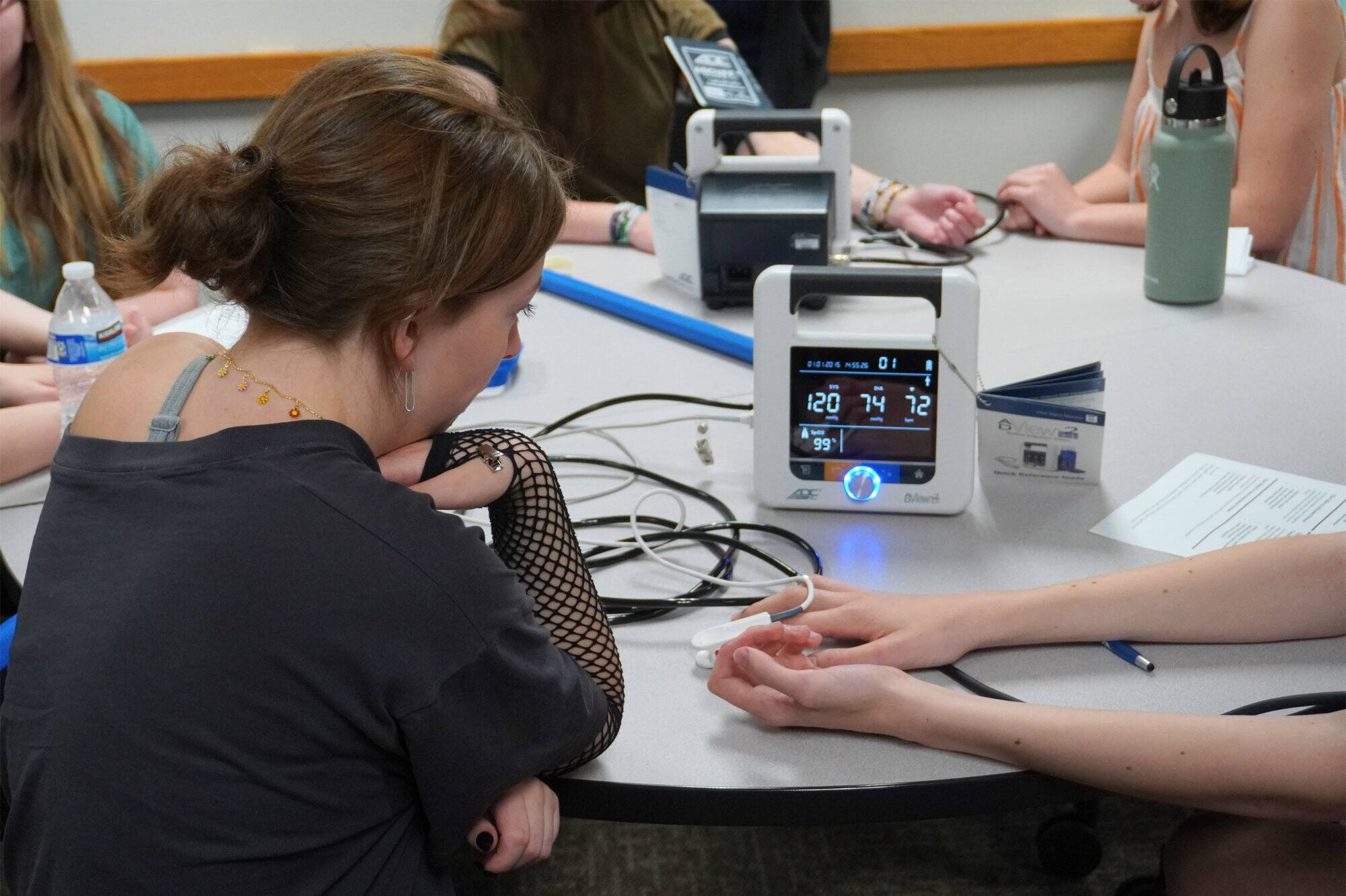 A student's blood pressure and pulse oximetry readings display on a medical monitor as a pulse oximeter is clipped to a classmate's finger, part of a hands-on healthcare or medical science activity around a shared table.